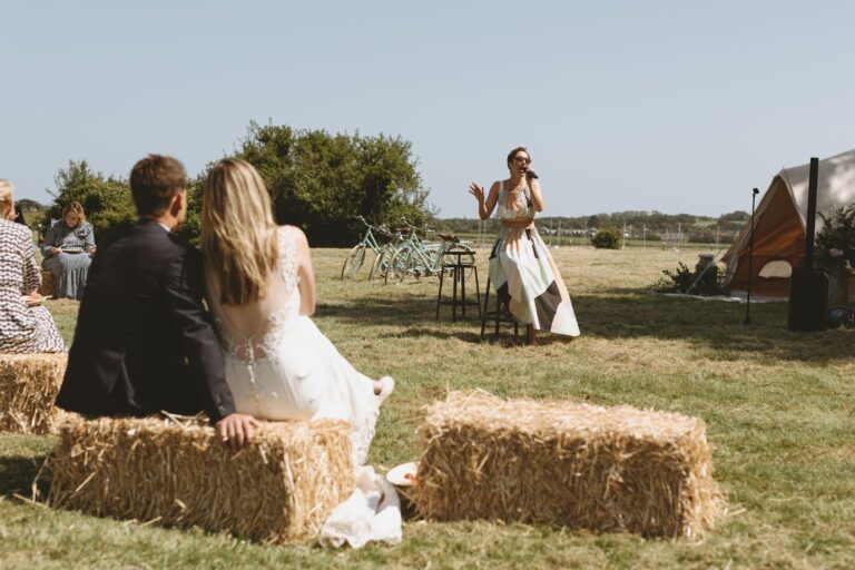 A singer performing on the lawn to a couple seated on straw bales, with a bell tent and bicycles behind, Pinkmead Estate