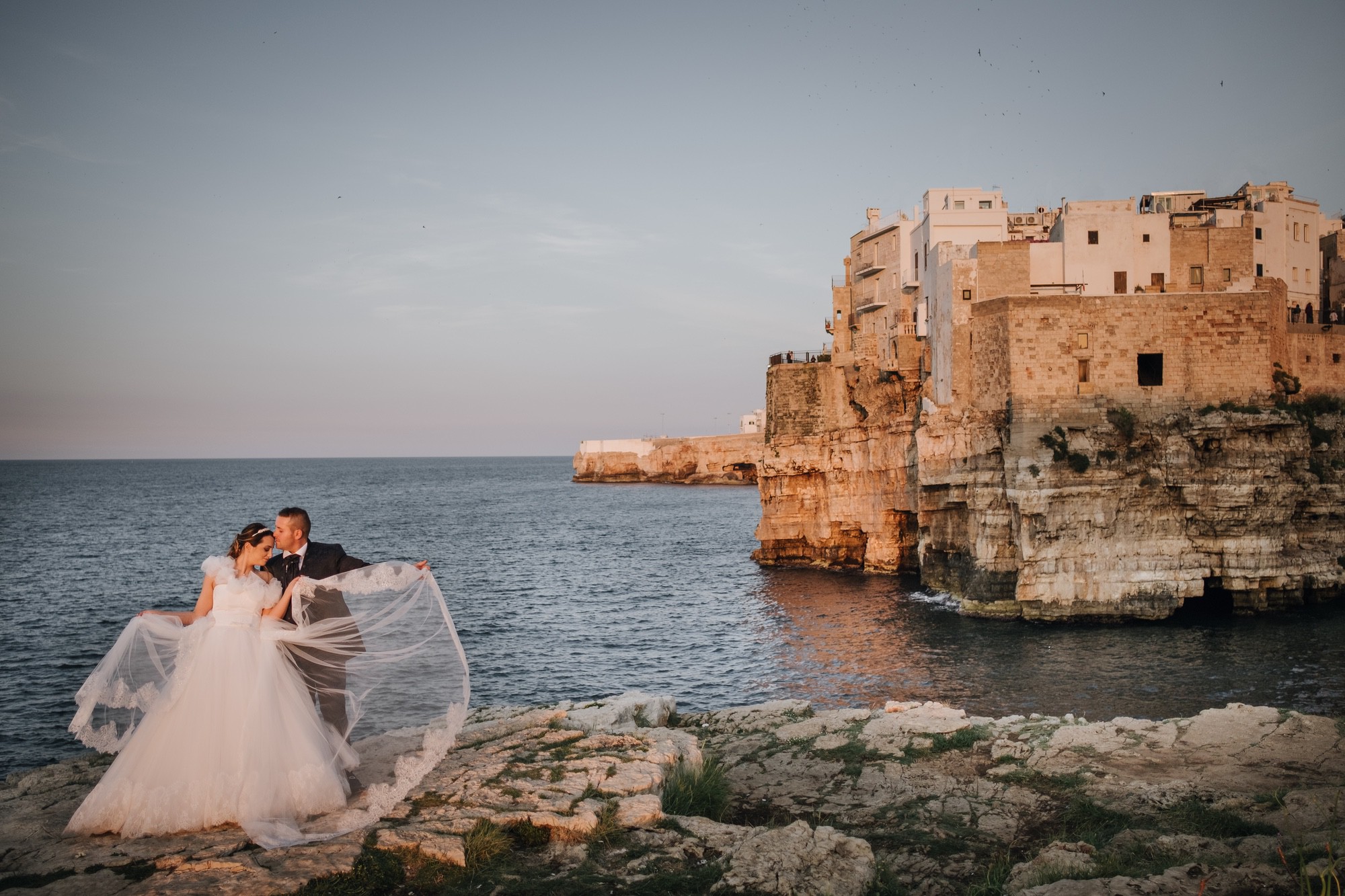 Bride and groom on the rocks of the Polignano a Mare seafront, the old town on its cliff behind them, Pino Coduti Photography