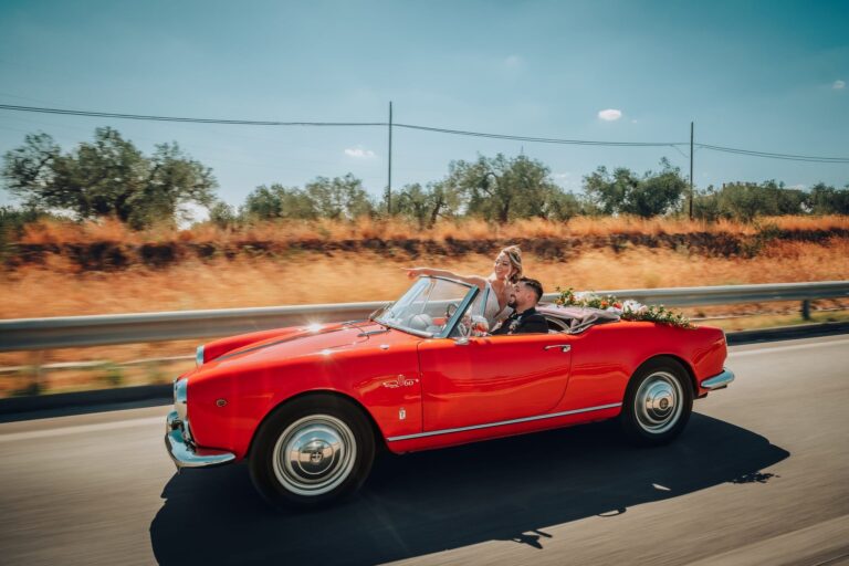 Bride and groom in a red open-top car, caught moving along a country road, Pino Coduti Photography