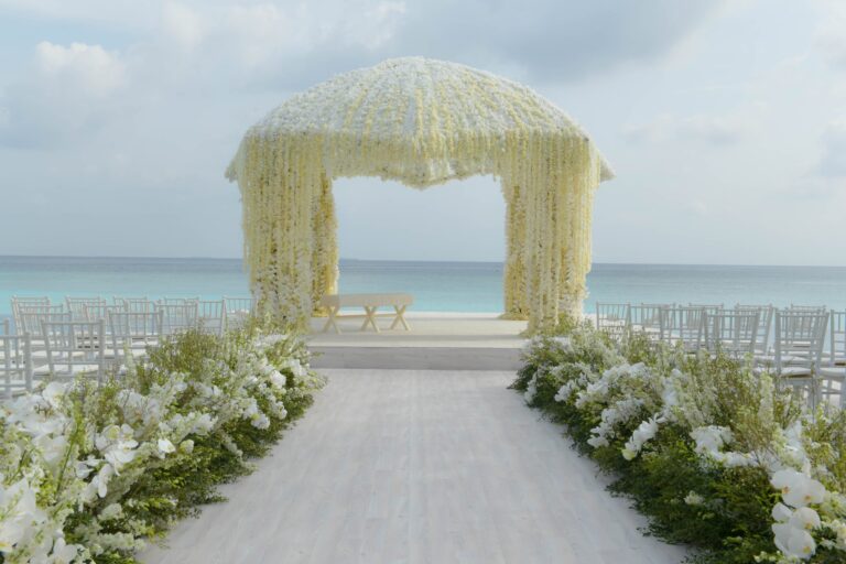 A white floral canopy and flower-lined aisle on a beach, set for a ceremony beside the sea, Preston Bailey
