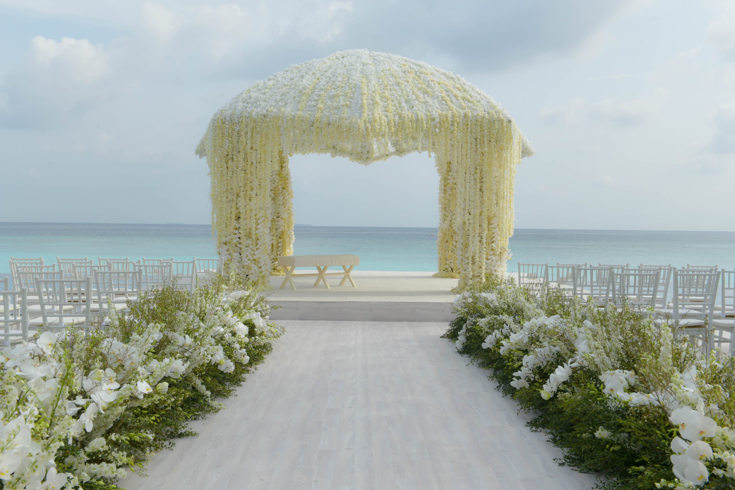 A white floral canopy and flower-lined aisle on a beach, set for a ceremony beside the sea, Preston Bailey