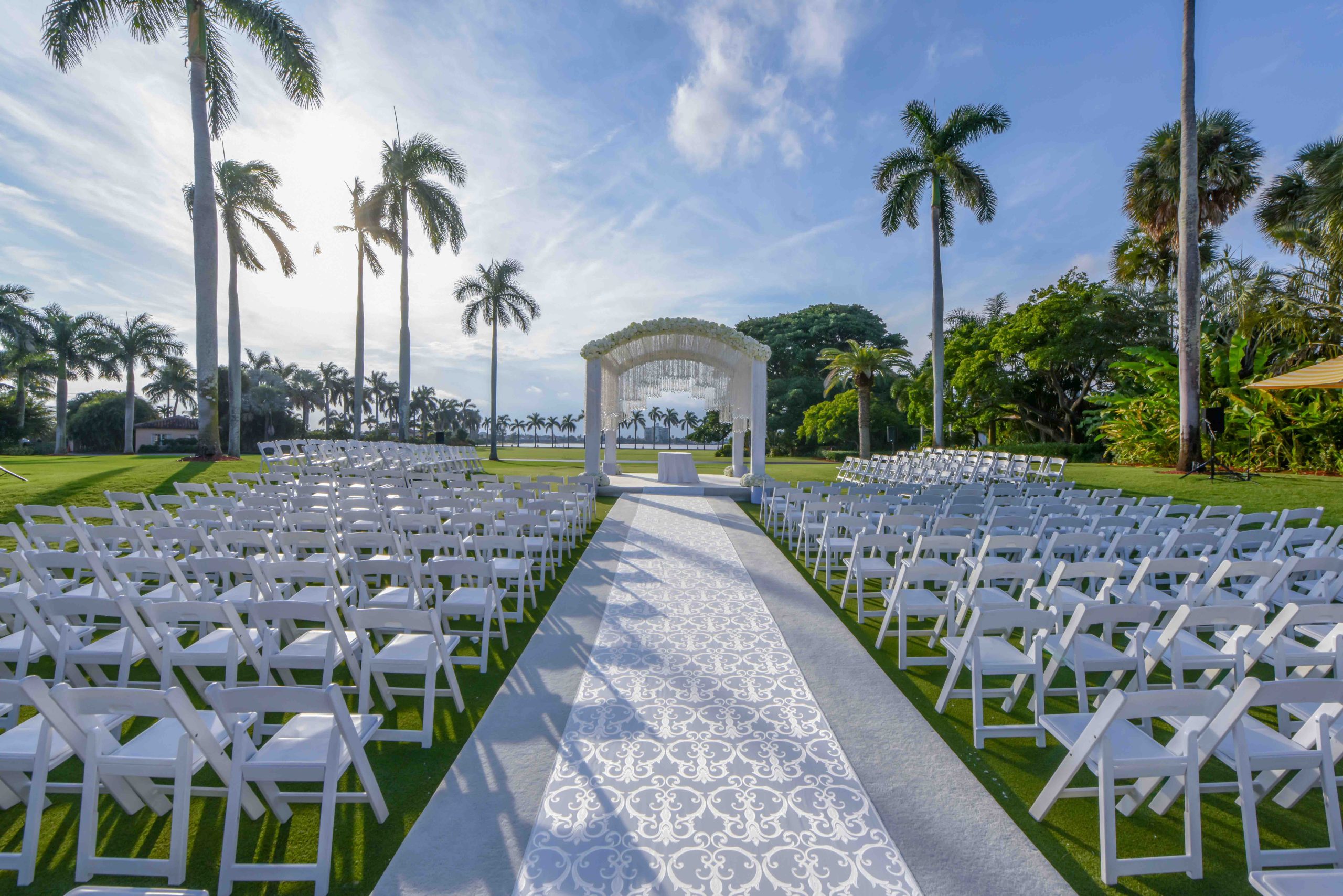 A white-and-lace ceremony aisle between rows of white chairs, leading to a pavilion among palm trees, Preston Bailey