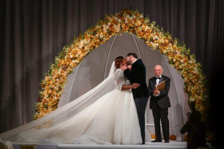 A couple kiss beneath an arch of orange and yellow flowers during a ceremony, Preston Bailey