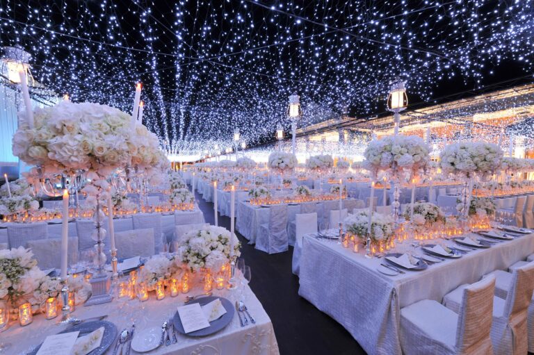 Long reception tables beneath a ceiling of fairy lights, with tall white floral centrepieces, Preston Bailey