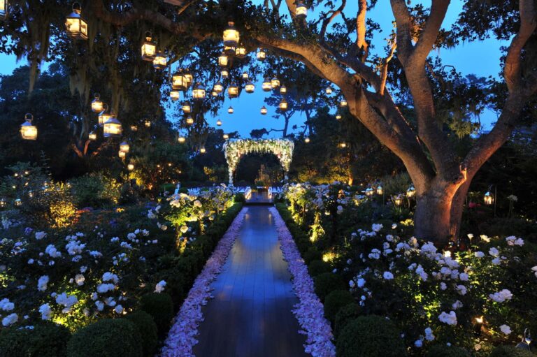 A garden lit by lanterns hanging from an old oak, with a reflecting channel leading to a floral arch, Preston Bailey