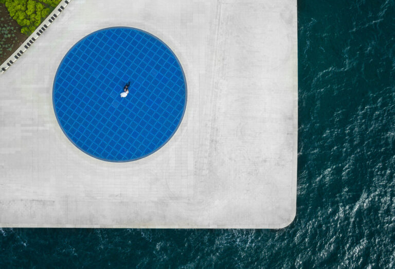 Aerial view of a couple on a blue circular pool beside the sea, Reflexion Studio