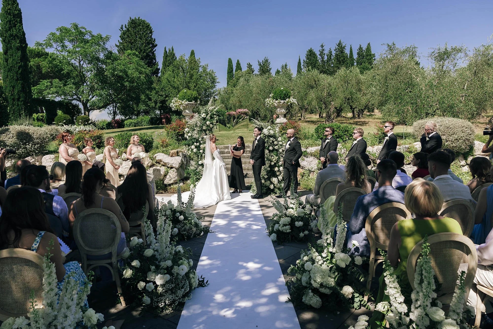 Bride and groom face each other as guests sit in the shade at a garden ceremony, Tenuta Corbinaia, Tuscany, Rellini Art Studio