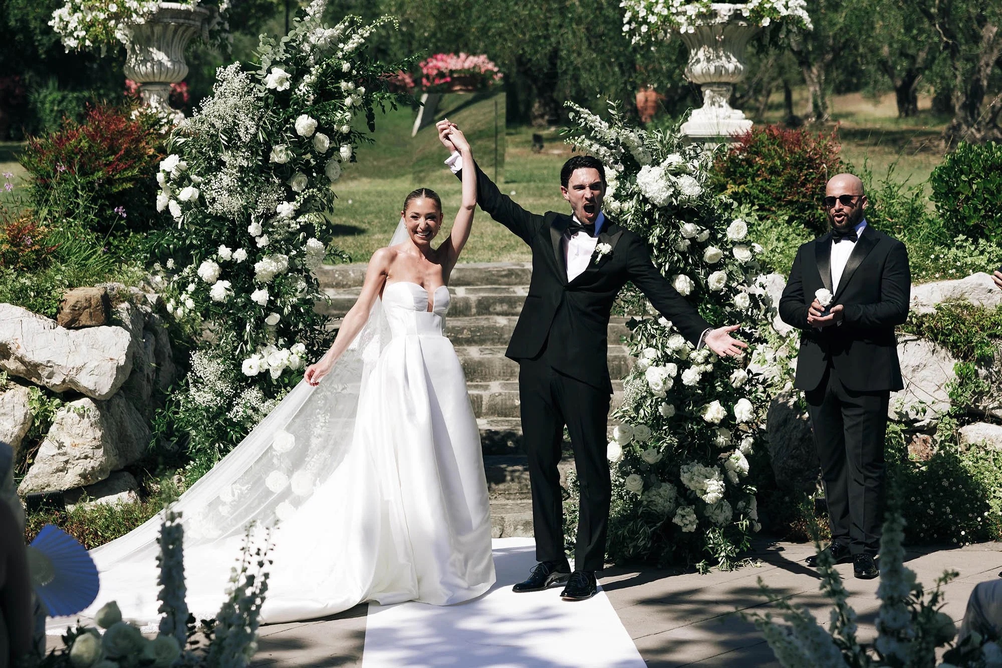 Arms in the air after the first kiss beneath a white floral arch, Tenuta Corbinaia, Tuscany, Rellini Art Studio