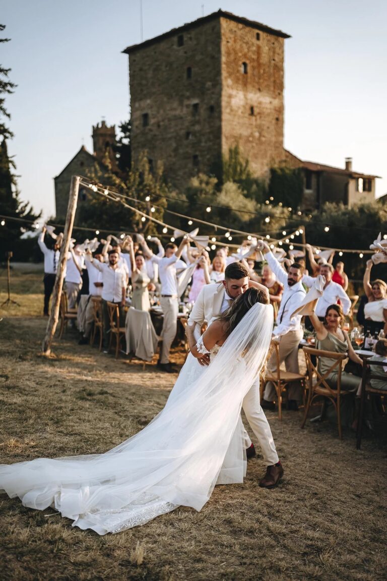 The couple kiss in front of their guests at an outdoor dinner below the tower of Castello di Ristonchi, Tuscany, Rellini Art Studio