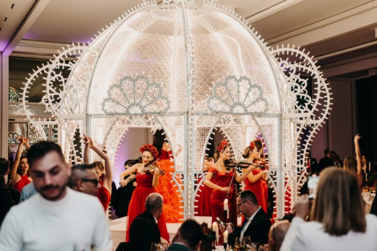 Violin ensemble performing under a lit white arch for seated dinner guests, Rinaldi Production