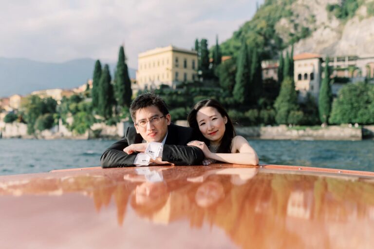 Florrie and Terrence resting on the deck of a wooden boat, a lakeside villa and cypresses behind them, during their Villa Cipressi wedding on Lake Como, Rocco Daniele Photography