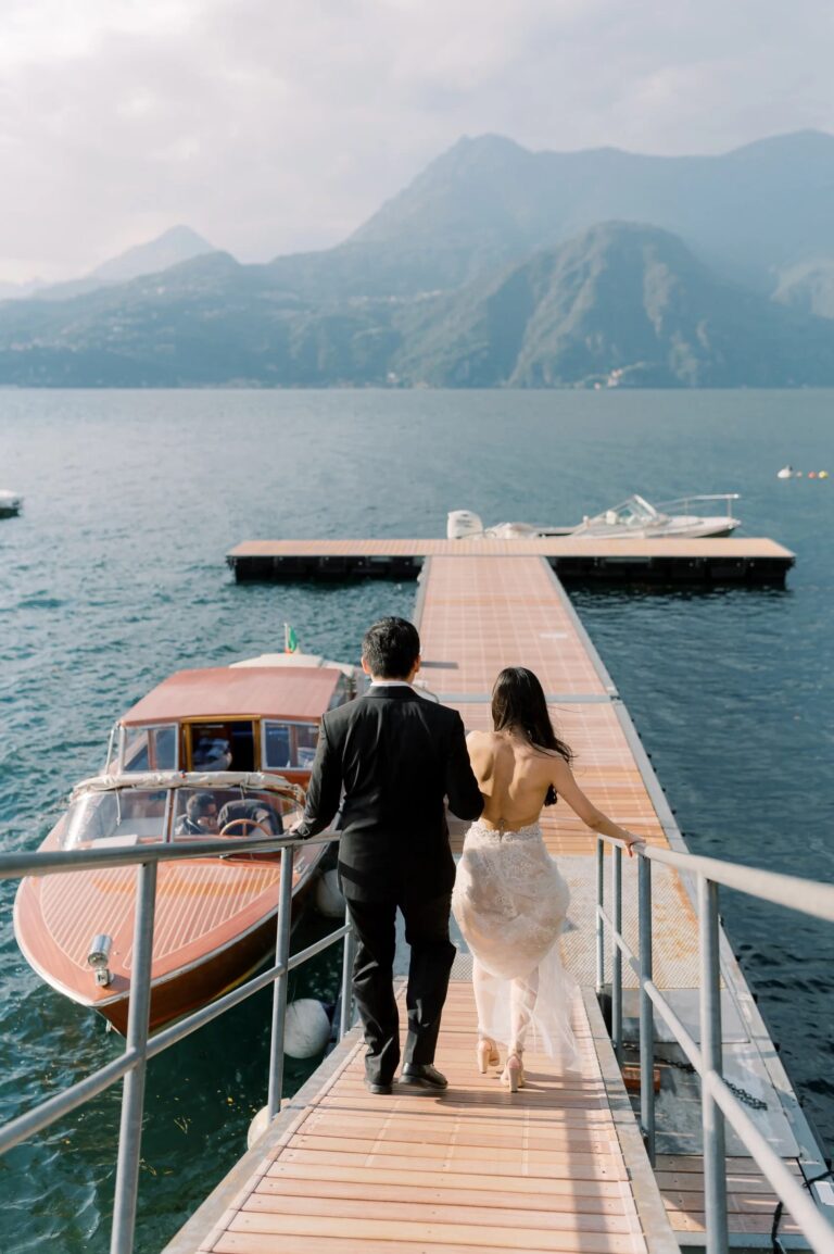 The couple walking down a jetty to their boat, mountains across Lake Como, Rocco Daniele Photography