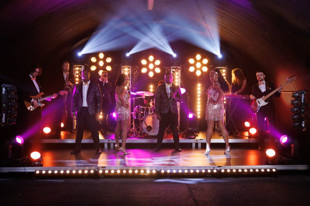 The full band on stage under spotlights and rows of lights