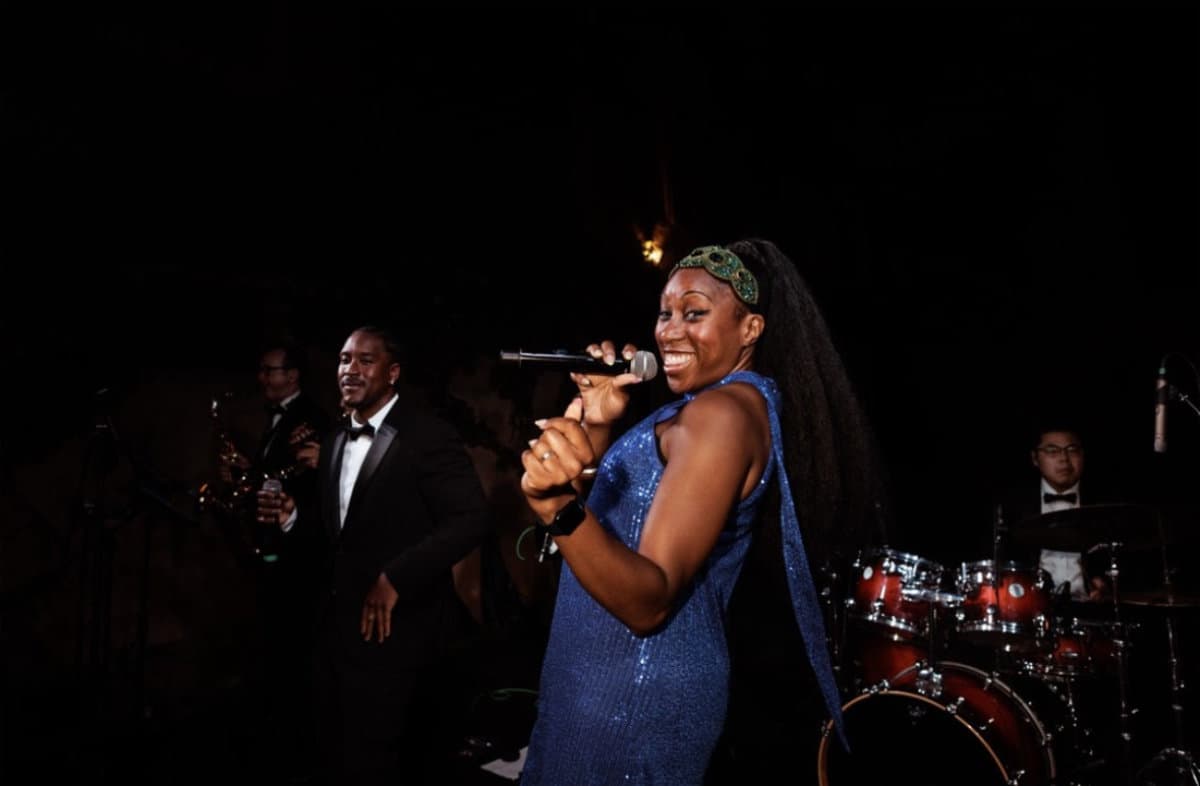 A vocalist in a blue dress and green headband singing, with the band in black tie behind her
