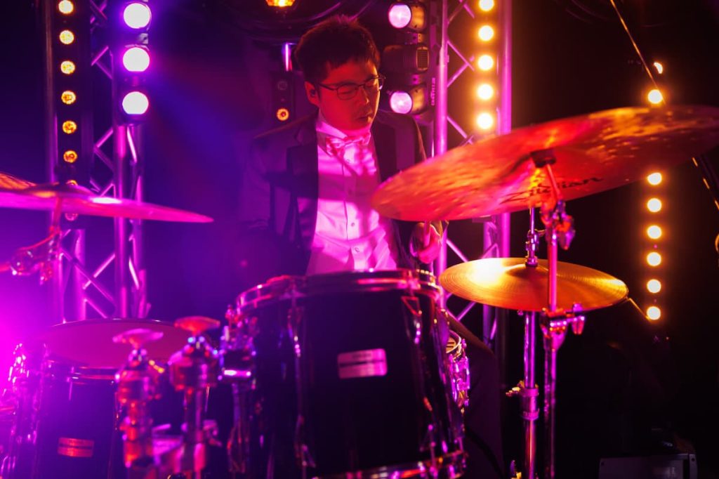 A drummer at a kit under pink and white stage lights