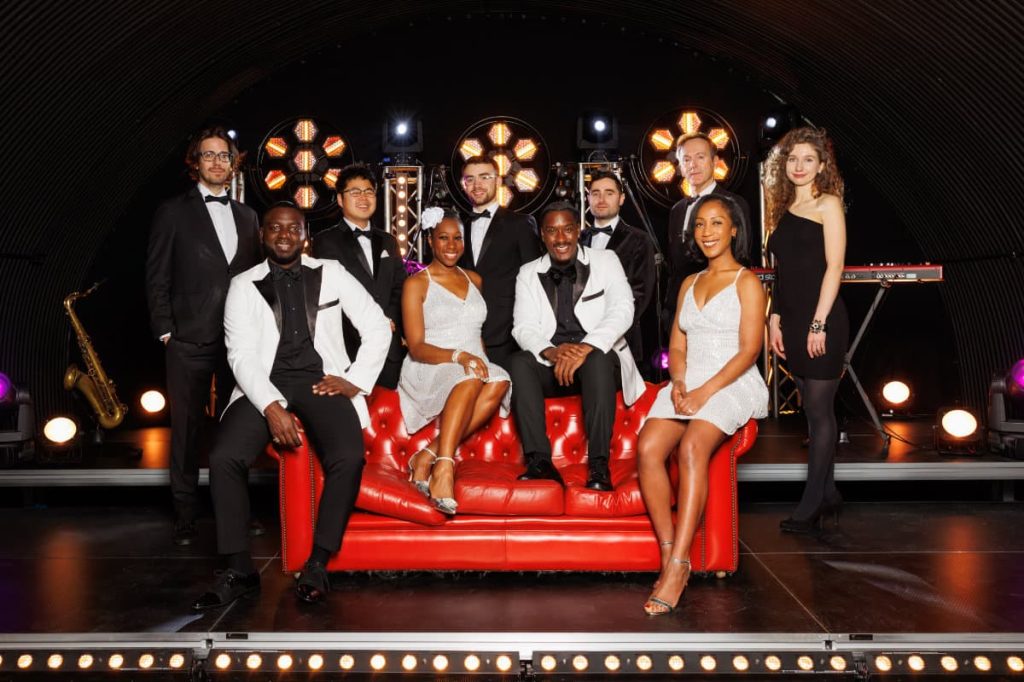 The band in black tie and evening dresses posed on a red sofa in front of stage lights