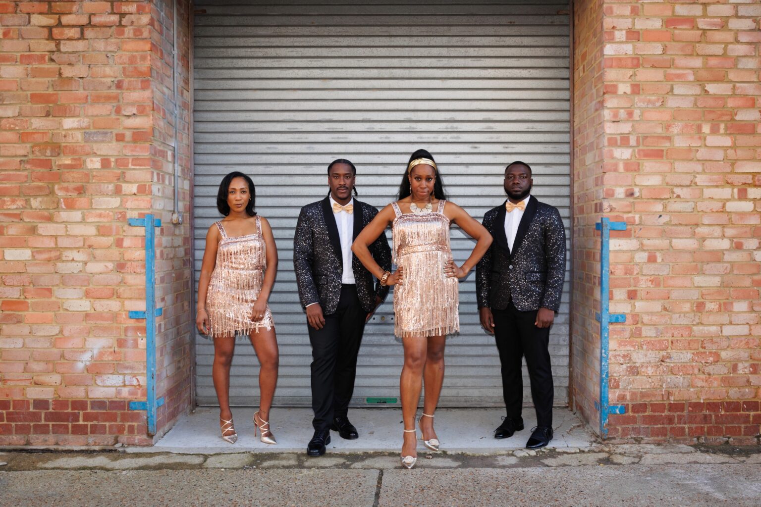 Four vocalists standing in front of a shuttered brick doorway in fringe dresses and sequin jackets
