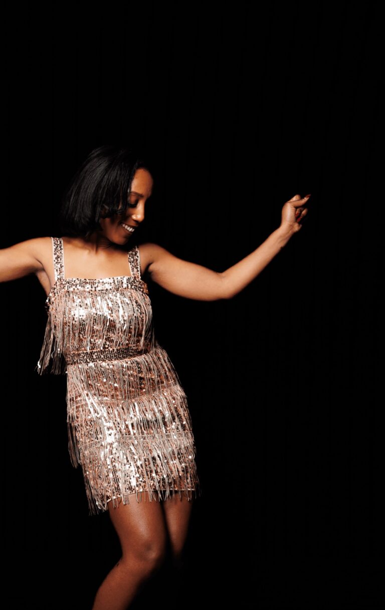 A vocalist dancing in a sequin fringe dress against a black backdrop, Rocks The Room