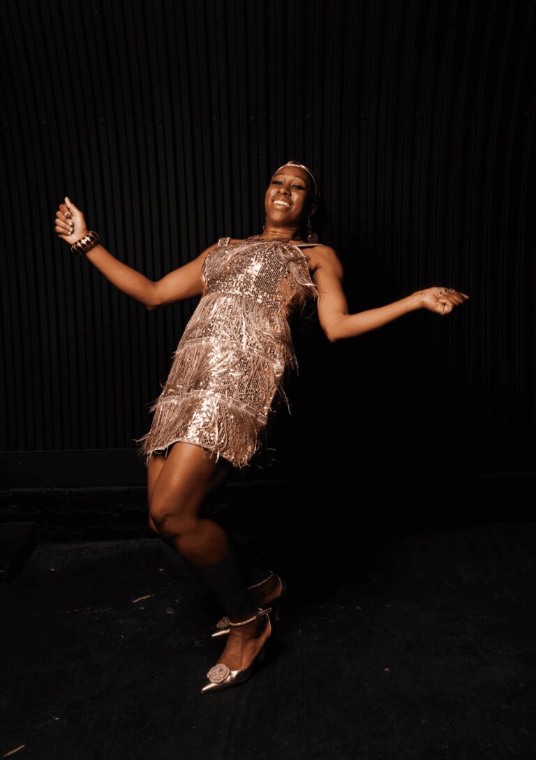 A vocalist dancing in a gold fringe dress with a headband against a black backdrop, Rocks The Room