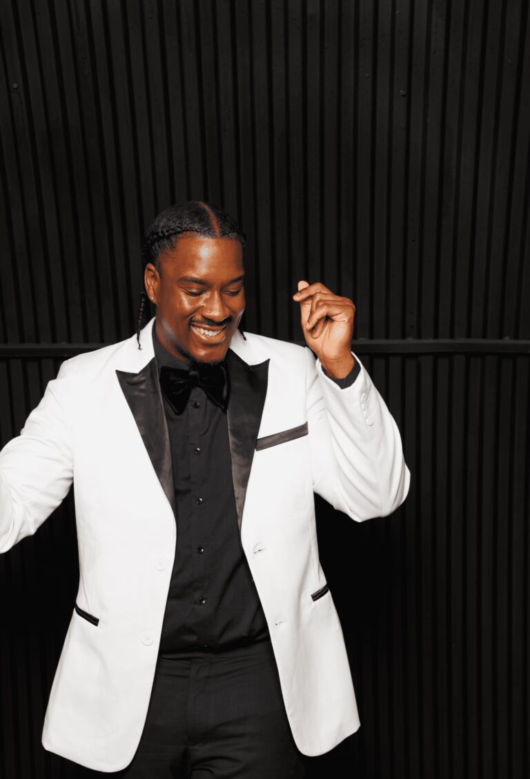 A male vocalist in a white dinner jacket smiling and dancing against a black backdrop, Rocks The Room