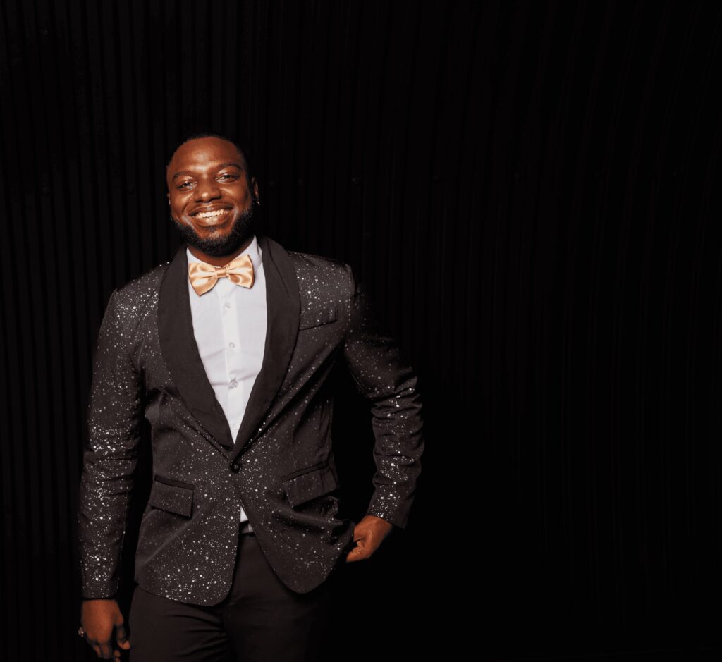 A male vocalist in a black sequin jacket and gold bow tie smiling in a dark studio, Rocks The Room