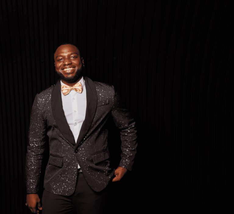 A male vocalist in a black sequin jacket and gold bow tie smiling in a dark studio, Rocks The Room