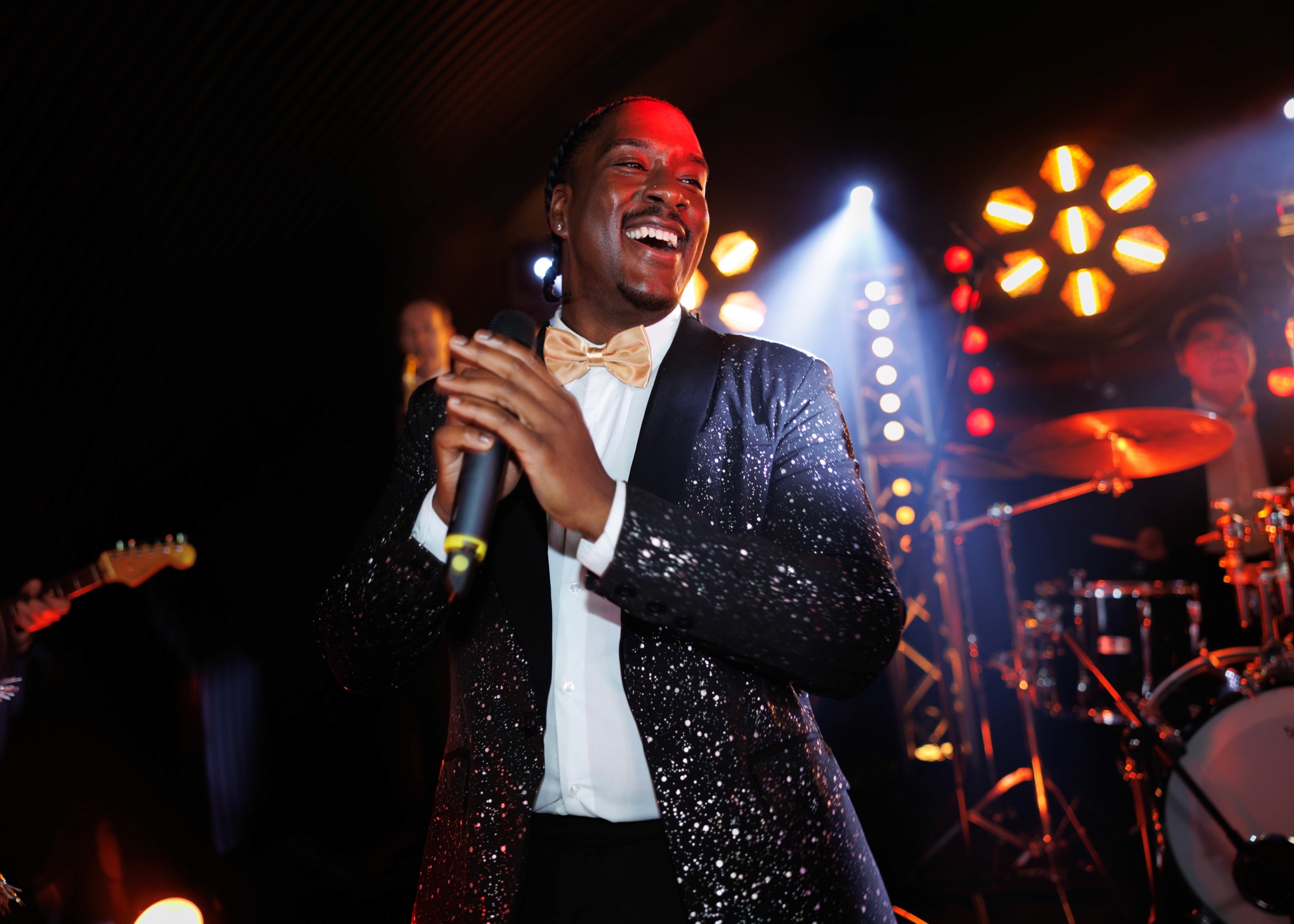 A male vocalist in a black sequin jacket and gold bow tie laughing mid-song with the band behind him, Rocks The Room