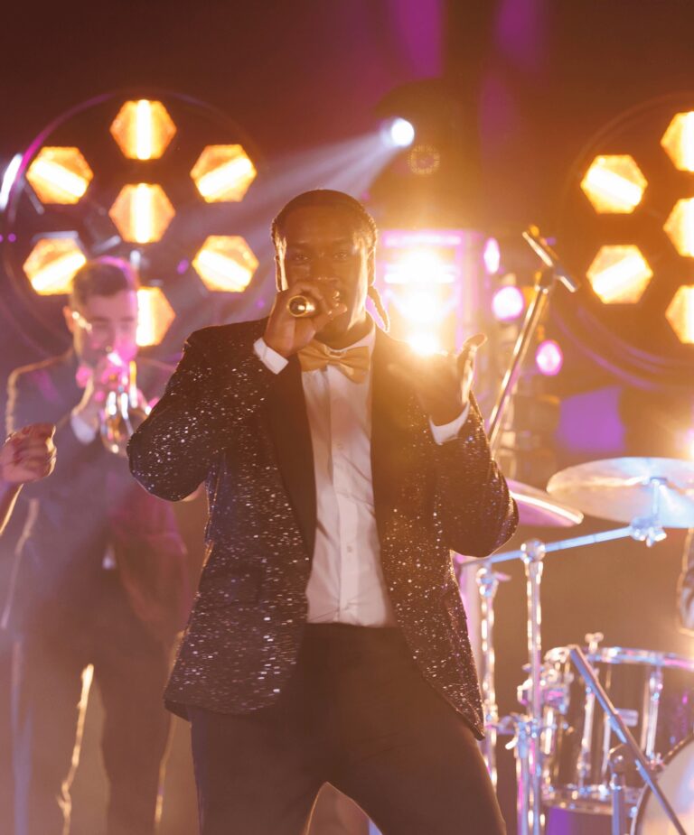 A male vocalist singing in glowing backlight with drums behind, Rocks The Room