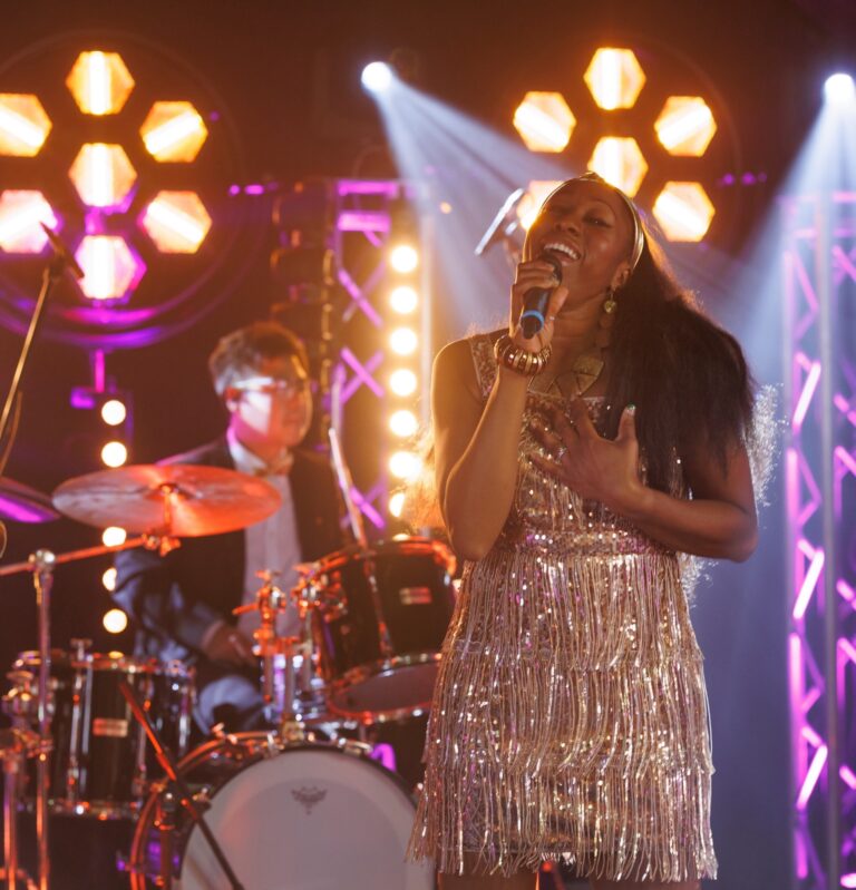 A female vocalist in a gold fringe dress singing, with the drummer behind her under gold lights, Rocks The Room
