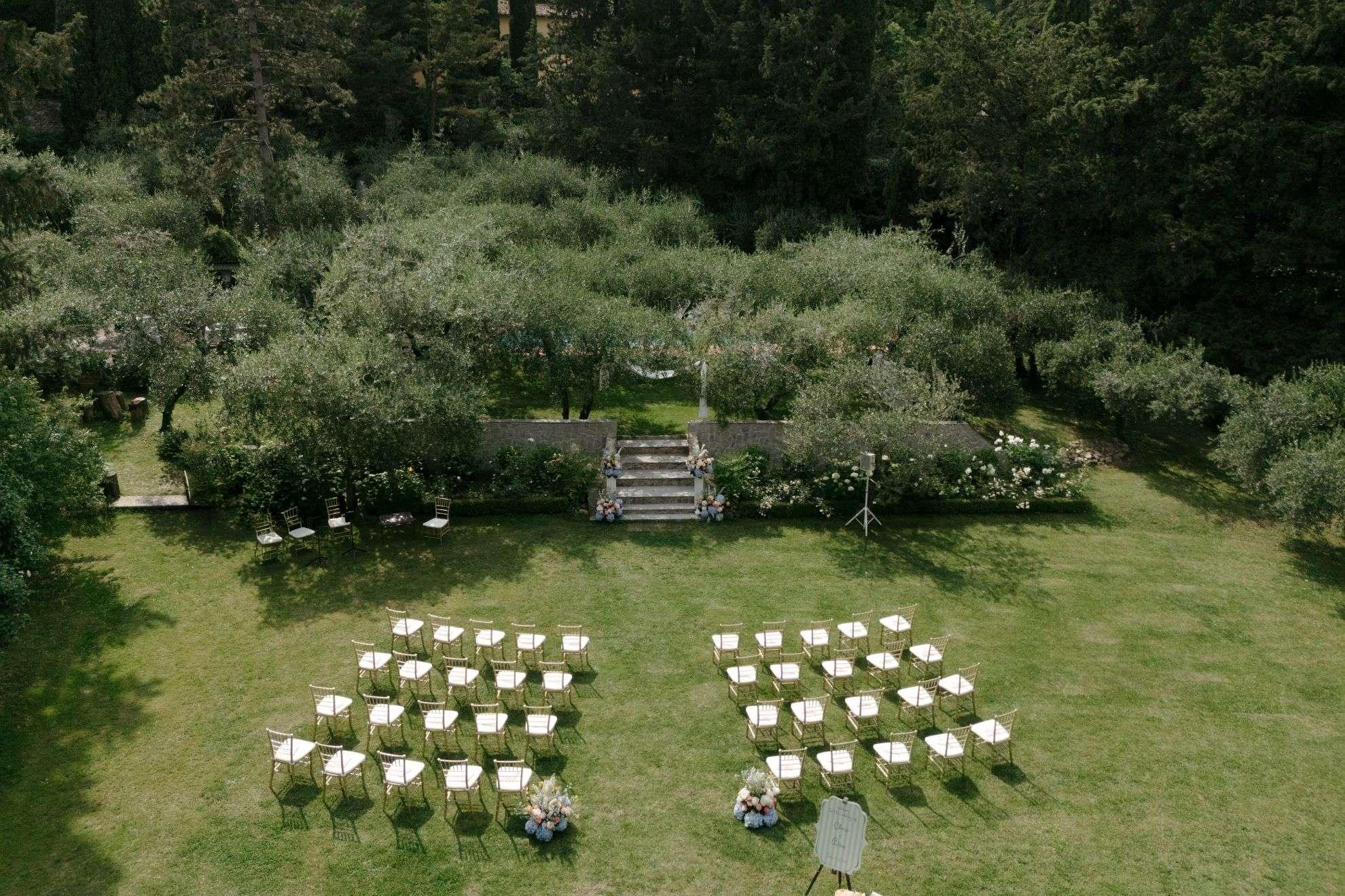 Aerial view of a ceremony set in a lawn garden among olive trees, white chairs either side of a stone stair (Chris & Ben), RoRas Destination Wedding