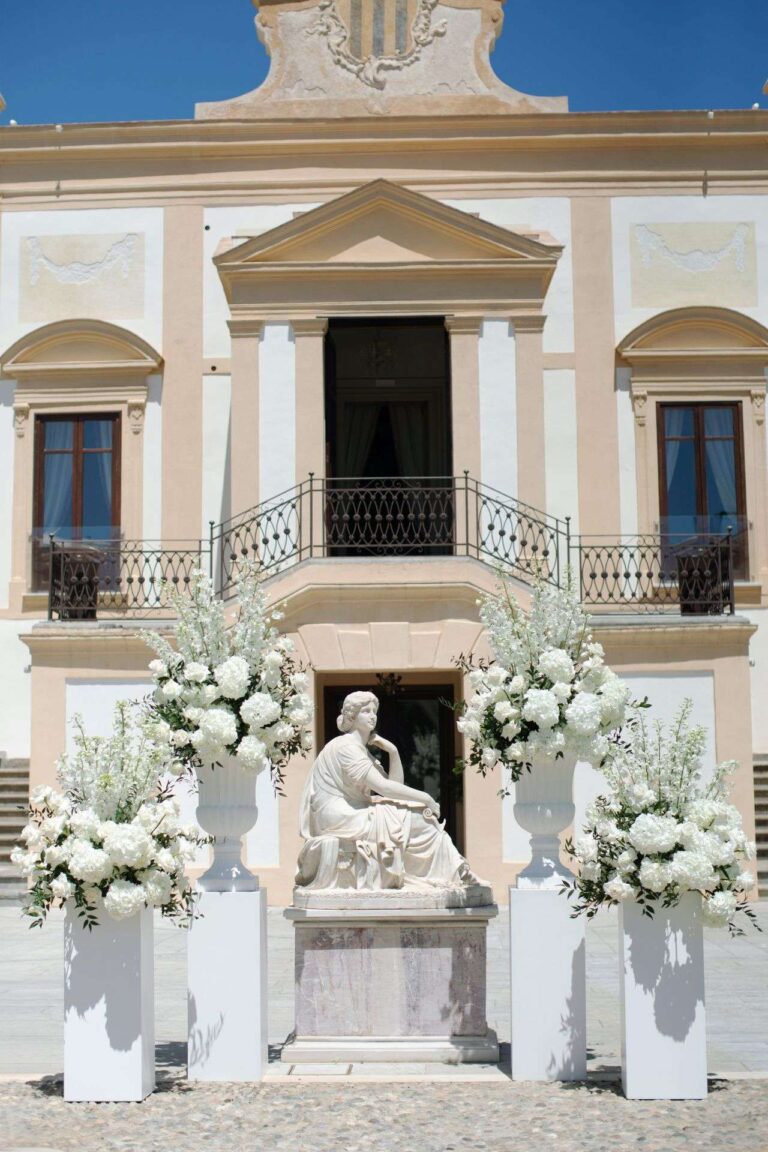 Ceremony set-up before a villa facade, white flowers on plinths either side of a marble statue (Ashley & Sergio; venue not named on the site), RoRas Destination Wedding