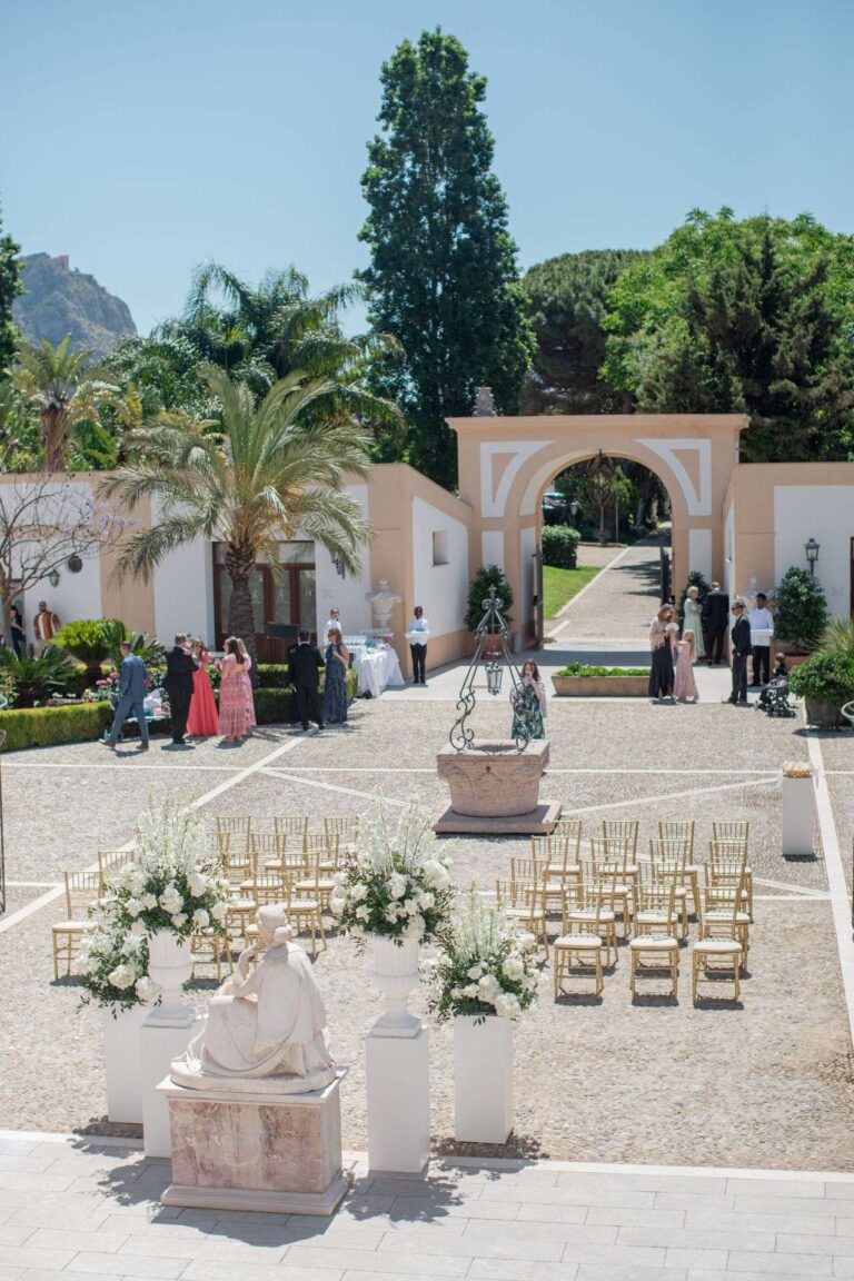 A courtyard laid out for the ceremony with gold chairs, white flowers and palms (Ashley & Sergio; venue not named on the site), RoRas Destination Wedding