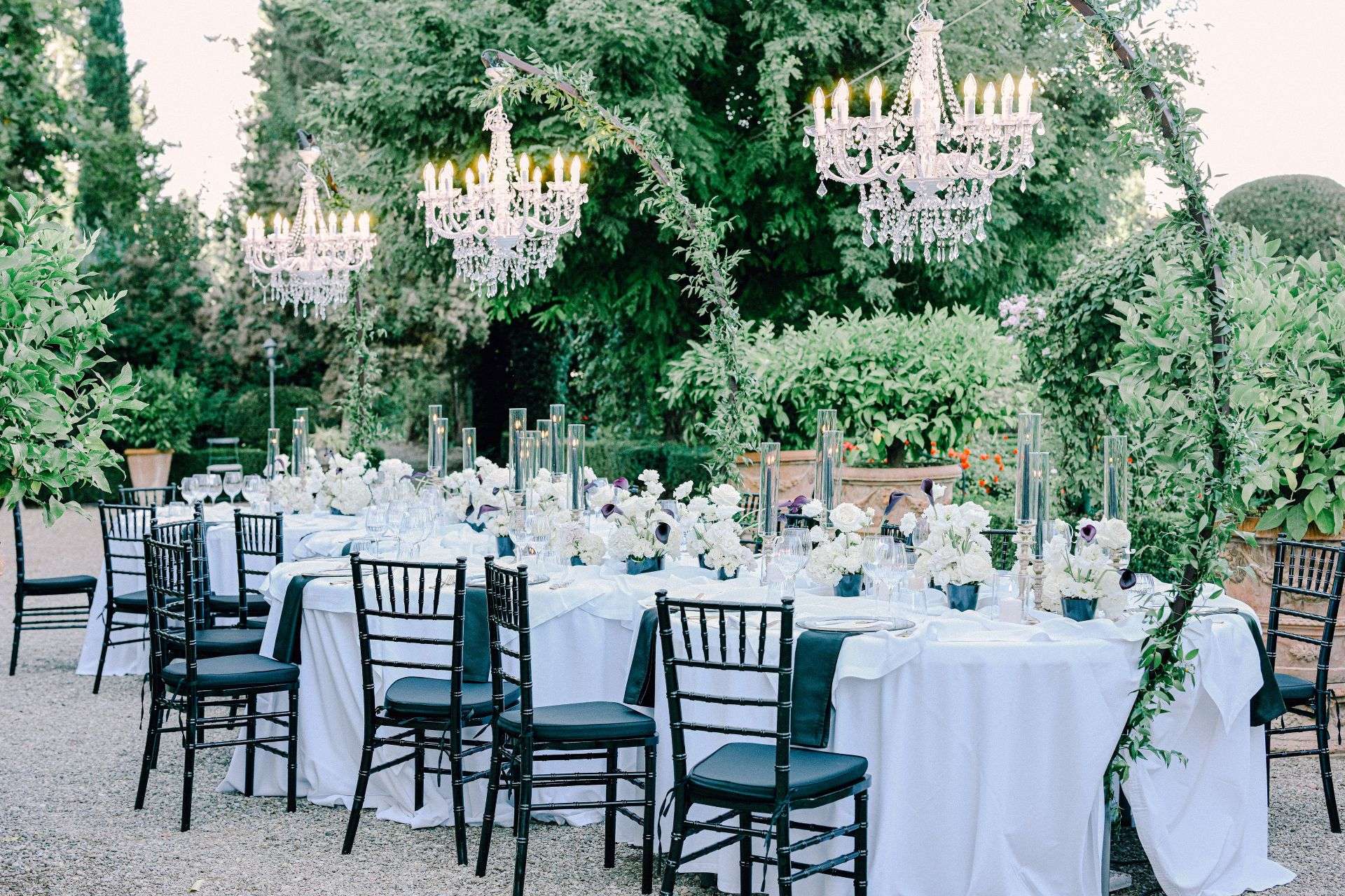 Reception tables set in a garden beneath hanging crystal chandeliers and a greenery arch, white linen and black chairs (Avery & Landon; venue not named on the site), RoRas Destination Wedding