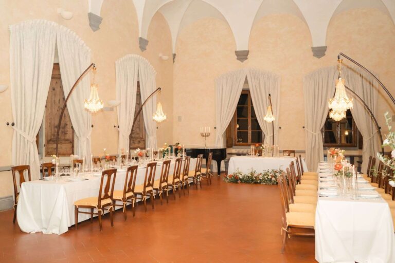 A vaulted hall with drapes and chandeliers set with long tables for dinner (Anna & Max; venue not named on the site), RoRas Destination Wedding