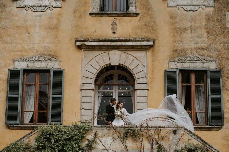The couple at an arched window of an ochre villa facade, the veil lifting in the wind, Villa di Ulignano, Tuscany (Sara & Marco), RoRas Destination Wedding