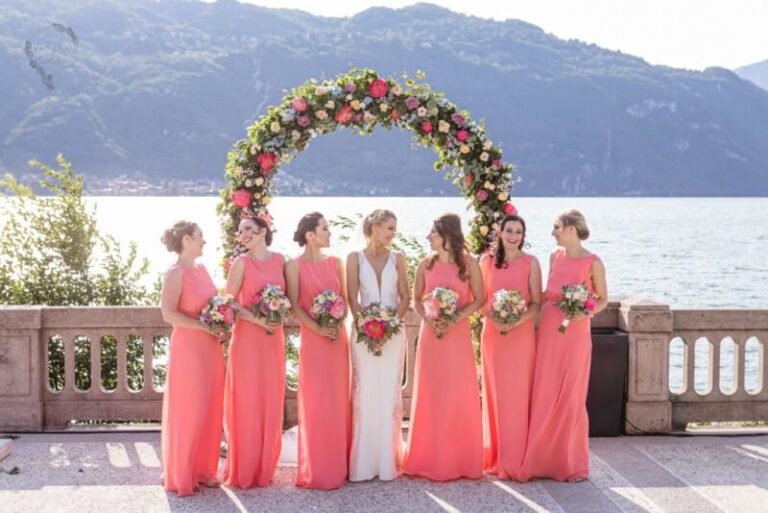 Bridal party in coral gowns beneath a floral arch on the lake shore, Samantha Pretto