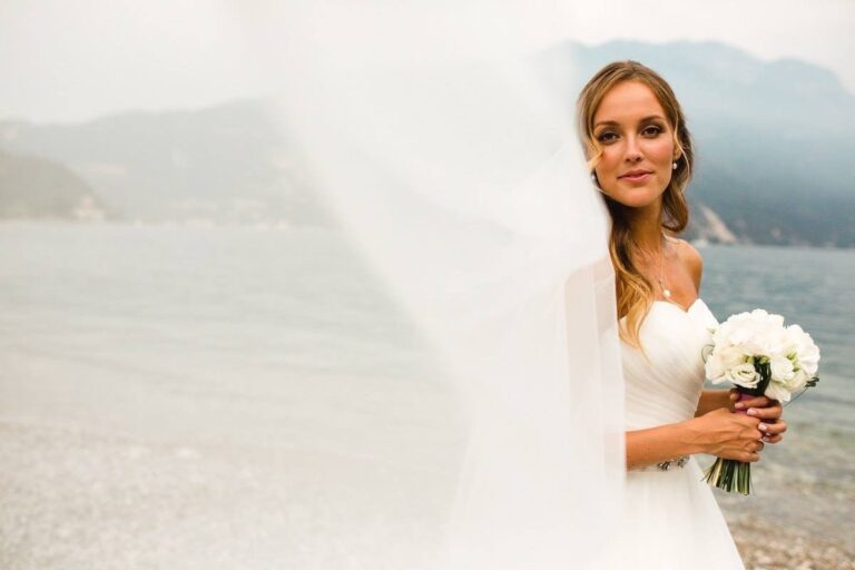 Bride with a veil lifted by the wind beside the lake, bouquet of white flowers, Samantha Pretto