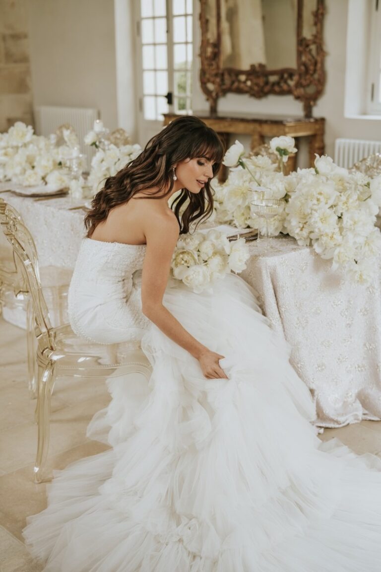 Bride arranging her gown beside a table of white floral arrangements, Paris, Sante Weddings