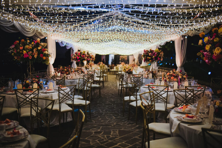 Round tables with red and pink flowers under strings of lights for an evening reception, Sara D'Angelo