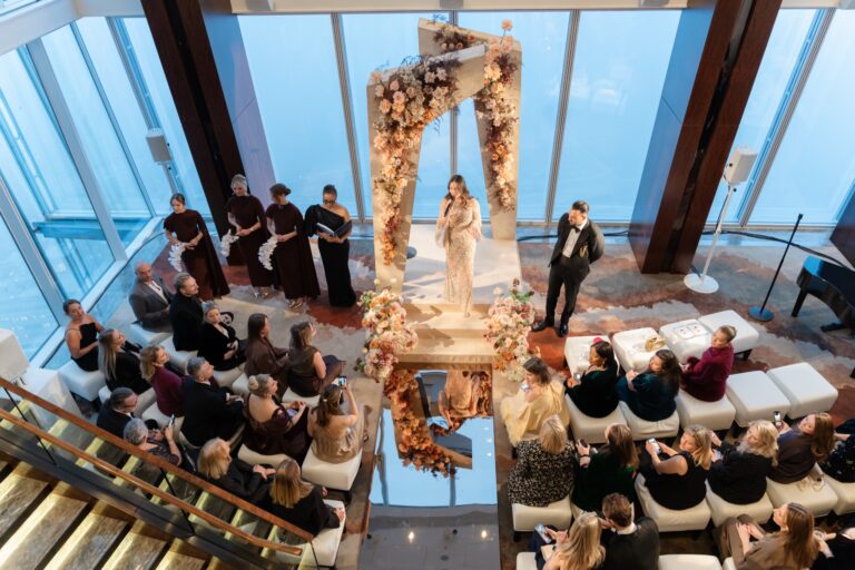A ceremony seen from the glass staircase, guests seated on white benches facing a floral arch against the windows, Shangri-La Hotel, The Shard, London