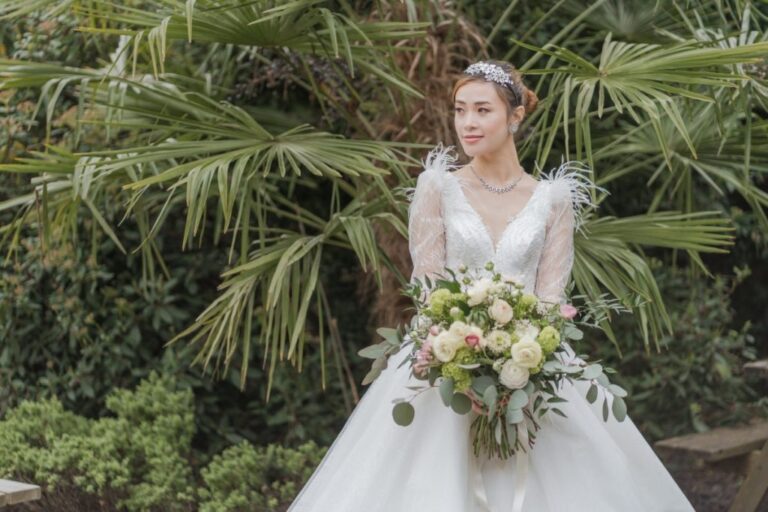 A bride in a feather-sleeve gown carries a bouquet of white rose and ranunculus, dressed with trailing eucalyptus, Shilpa Reddy