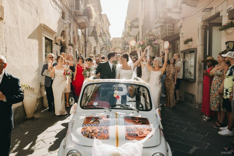 A white Fiat 500 dressed for the wedding in a narrow street, guests applauding, Sicilian Wedding Day