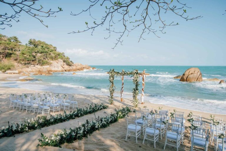 Beach ceremony set with a driftwood arch and pale blue chairs above a flower-lined aisle, Silavadee Pool Spa Resort
