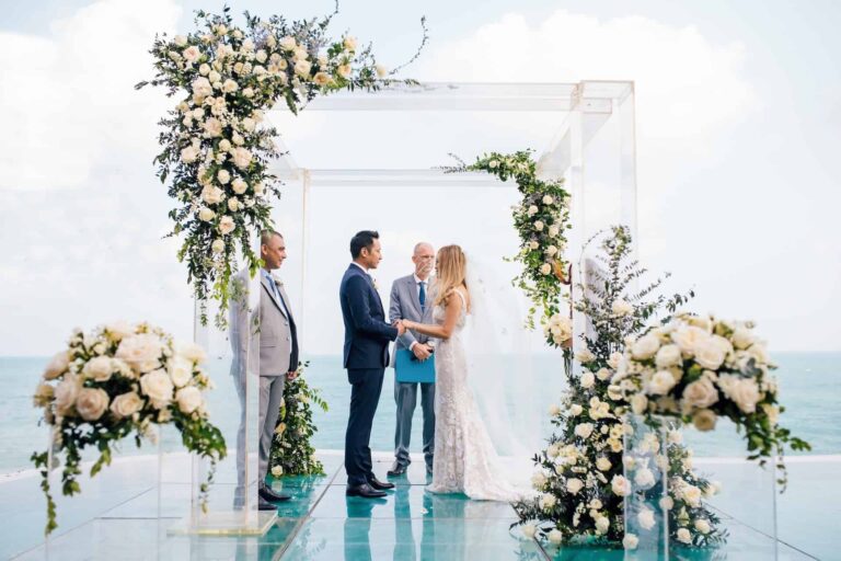 Vows beneath a white floral arch on a glass platform with the sea behind, Silavadee Pool Spa Resort