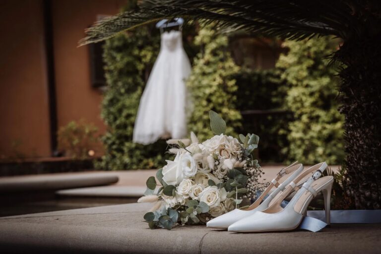 The dress hangs beneath a palm while the bouquet and shoes wait on a stone ledge, Simone Perini Photography