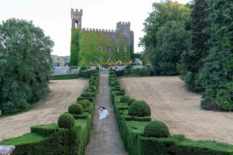 A couple small at the foot of a long box-hedged avenue below an ivy-covered castle, Simone Perini Photography