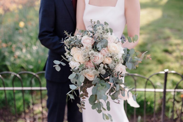 A bride and groom with a bouquet of roses and eucalyptus beside park railings, Sofitel London St James