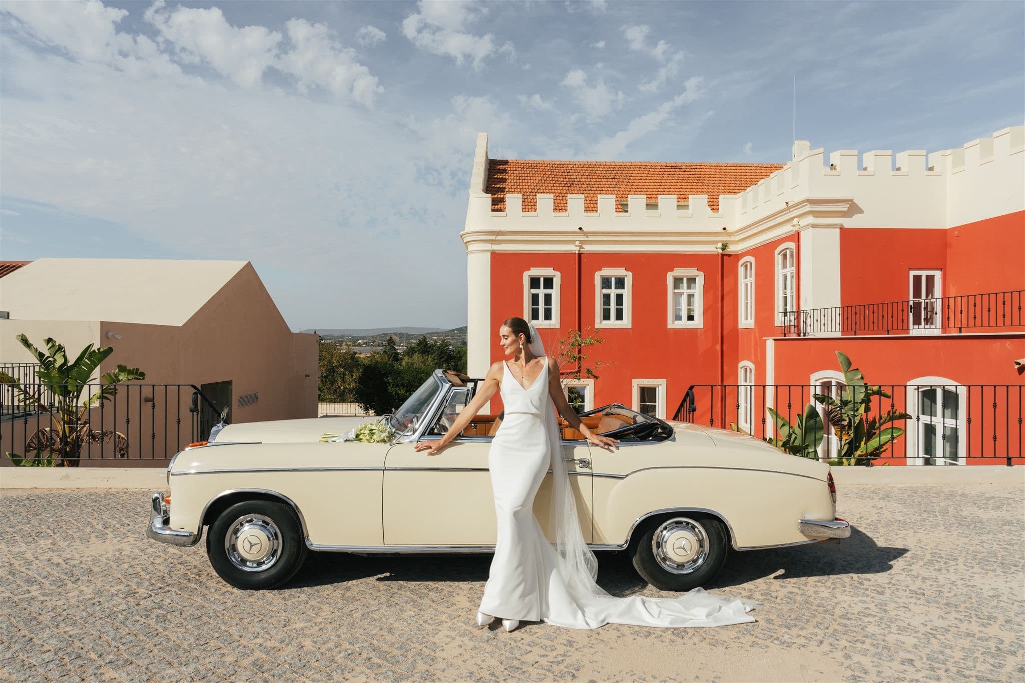 A bride in a white dress beside a cream vintage convertible in front of the red Palace, Solar do Paço Algarve