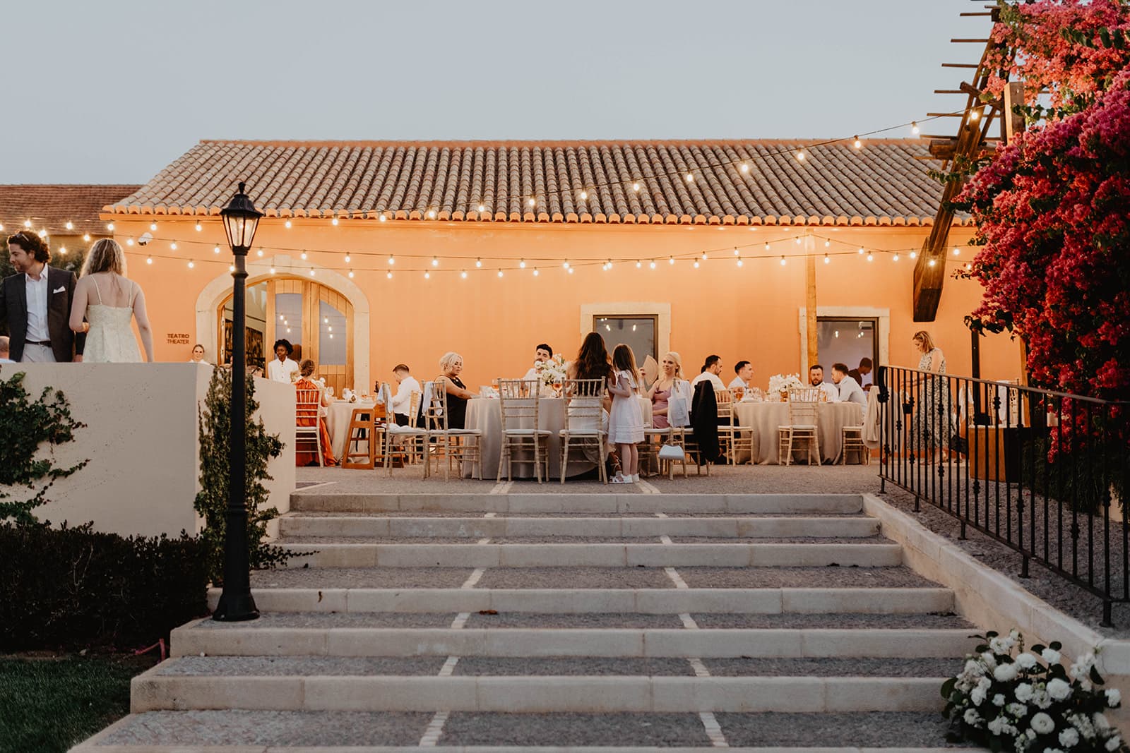Guests at round tables on a terrace at dusk beneath strings of lights, with stone steps in front, Solar do Paço Algarve