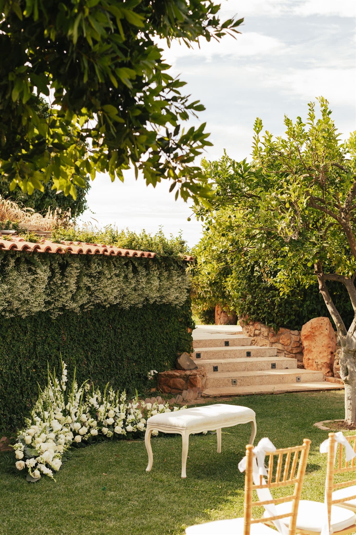 A white bench and white flowers at the foot of garden steps beside chairs set for a ceremony, Solar do Paço Algarve